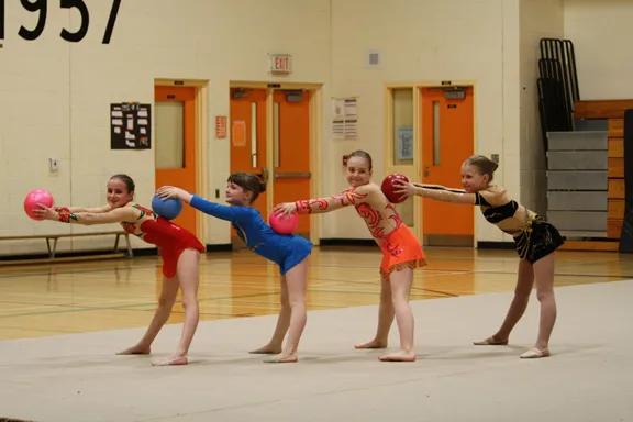 A line of gymnasts in bright leotards stretching forward with balls.