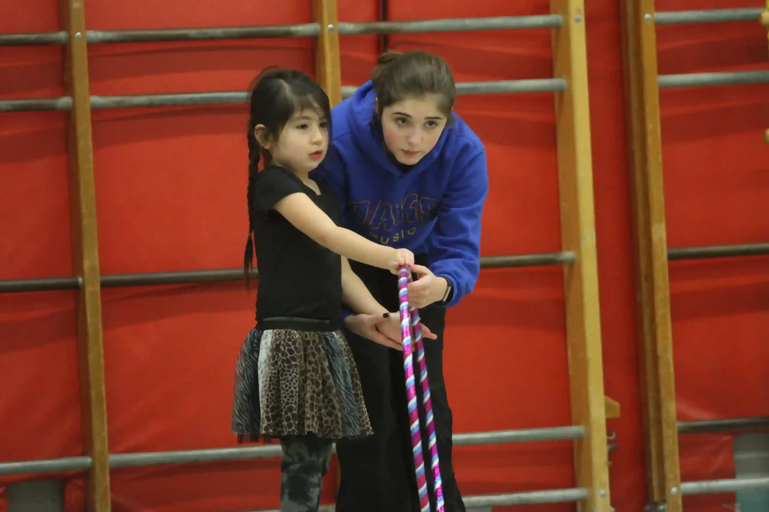 A coach in a blue club hoodie leaning in to help a young gymnast hold her hoop.